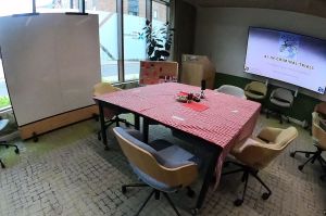 Empty chairs around a table with a red tablecloth and pens and paper, screen in background
